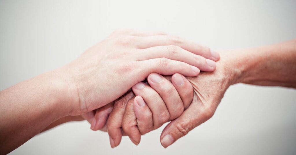 Two hands clasped in a supportive grip, with a younger hand over an older hand to show care and support.