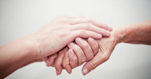 Two hands clasped in a supportive grip, with a younger hand over an older hand to show care and support.