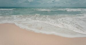 Turquoise ocean waves wash onto a pale sandy beach under a partly cloudy sky.
