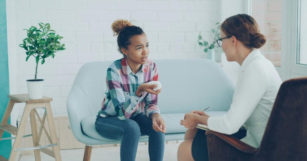 image - Treat Mental Health California A young woman on a light blue couch talks with a counselor in a bright room, holding a stress ball.