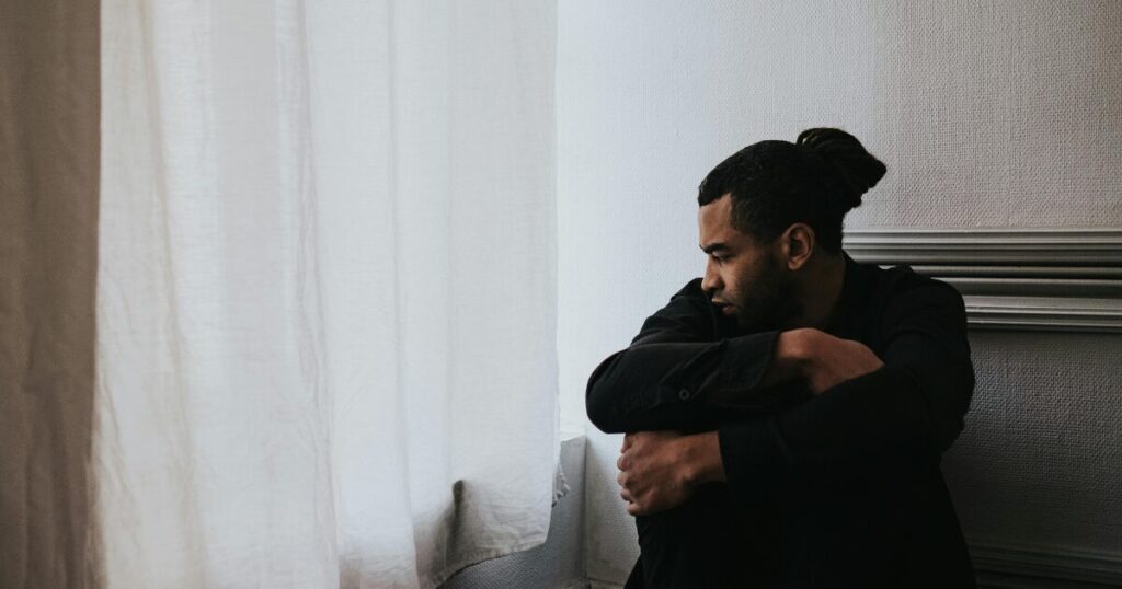 image - Treat Mental Health California Man sitting on the floor by a window with sheer curtains, arms wrapped around his knees, looking down in a contemplative mood.