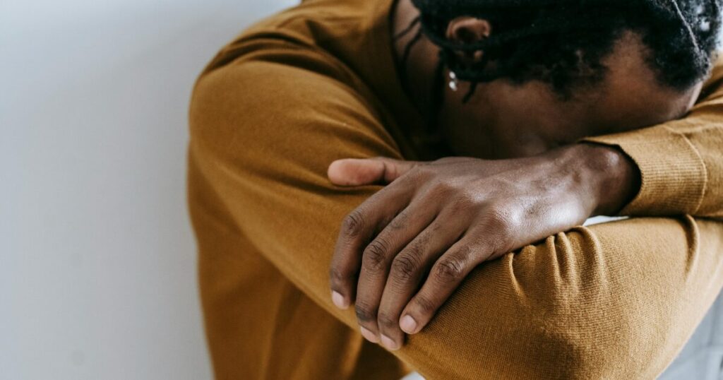image - Treat Mental Health California Person sits with their head bowed onto crossed arms, wearing a brown sweater, suggesting sadness or fatigue.