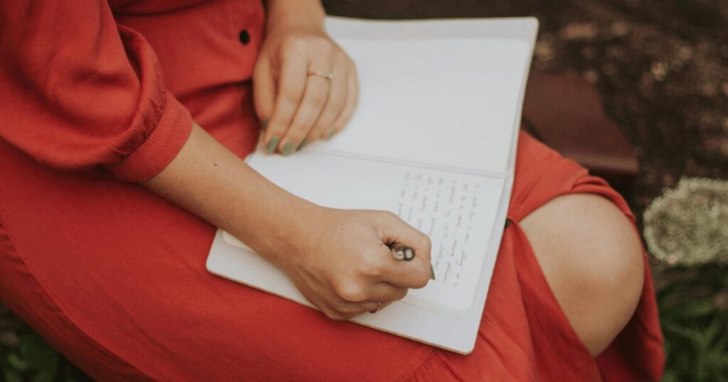 Person in a red dress writes in a white notebook resting on their lap outdoors.