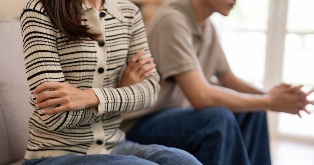 Two people sit on a sofa; foreground person in a striped cardigan has arms crossed, while the other sits nearby with hands clasped.