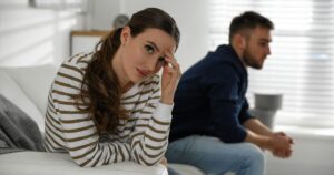- Treat Mental Health California Woman in a white-and-brown striped sweater sits on a white sofa, touching her forehead with a worried expression as a man in a dark hoodie sits apart in the background in a bright living room.