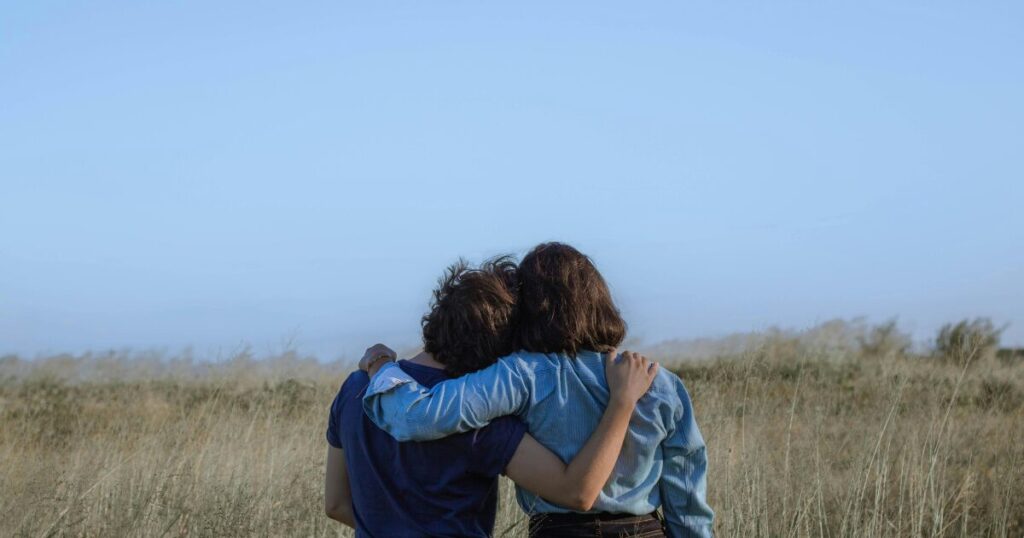Two people with their backs to the camera, arms around each other, standing in a tall golden field under a clear blue sky.