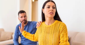 - Treat Mental Health California Woman in a yellow knit sweater looks away as a man in a blue shirt places a hand on her shoulder in a living room, appearing tense or concerned.