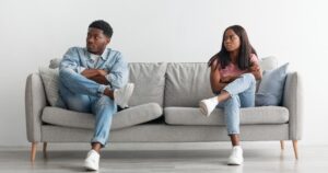 Two people sit on a light gray sofa facing away from each other with arms crossed, looking unhappy.