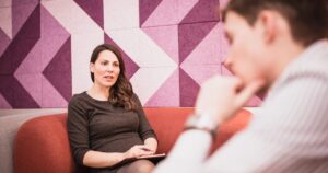 - Treat Mental Health California Woman seated on an orange couch talks to a blurred man in the foreground, with a pink geometric wall behind them.