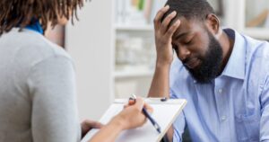 - Treat Mental Health California Man with a beard sits with head in hand, expressing distress during a counseling session with a clinician taking notes on a clipboard.