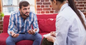 - Treat Mental Health California Man discusses concerns with a therapist seated on a red couch during a counseling session in a brick-walled room.