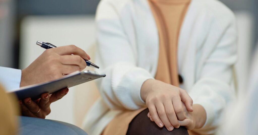 Person writes on a clipboard with a pen while another person sits with folded hands in a calm setting.