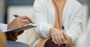 - Treat Mental Health California Person writes on a clipboard with a pen while another person sits with folded hands in a calm setting.