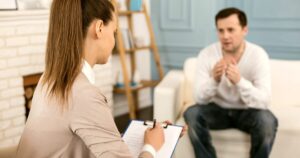 - Treat Mental Health California Therapist taking notes on a clipboard as a man talks, seated on a sofa in a cozy therapy room.