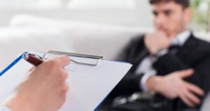 - Treat Mental Health California Therapist taking notes on a clipboard while a man in a suit sits on a couch, suggesting a counseling session.