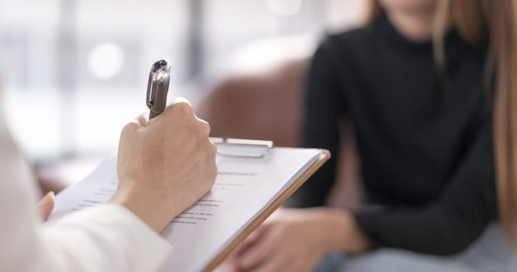 Person writes on a clipboard with a silver pen during a consultation, blurred figure in a dark top in the background