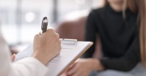 Person writes on a clipboard with a silver pen during a consultation, blurred figure in a dark top in the background