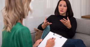 Woman in a black top sits on a sofa, explaining with hand gestures while a counselor takes notes on a clipboard nearby.