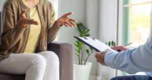 Counselor takes notes on a clipboard while client speaks and gestures with open hands in a therapy session.