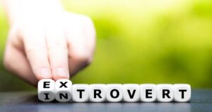 Hand arranging dice that spell 'EX INTROVERT' on a table with a green blurred background.