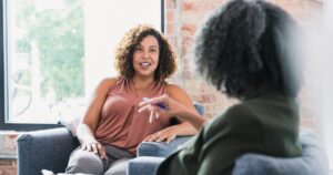 Two women chat on a blue sofa near a brick wall in a bright room.