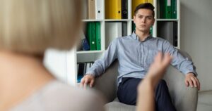 Man in a light blue shirt sits in an armchair, talking with a therapist in a tidy office. Shelves with binders are in the background, a blurred figure in the foreground suggests a session.