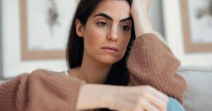 Woman with long dark hair sits indoors, resting her head on her hand with a thoughtful, sad expression.