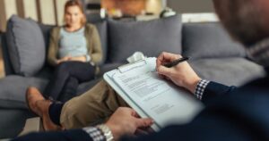 Therapist writing notes on a clipboard while a client sits on a sofa in a cozy living-room setting.