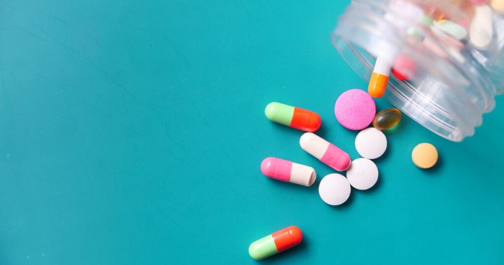Assorted pills and capsules spilled from a clear bottle onto a turquoise background.