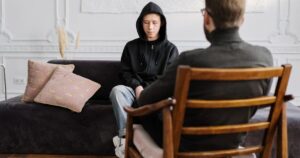 Teenager in a black hoodie sits on a dark sofa with pink pillows, facing a seated therapist in a wooden chair.