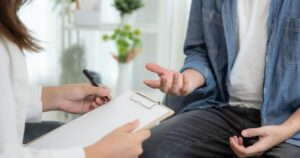 - Treat Mental Health California Therapist taking notes on a clipboard while a client gestures and talks in a calm counseling room