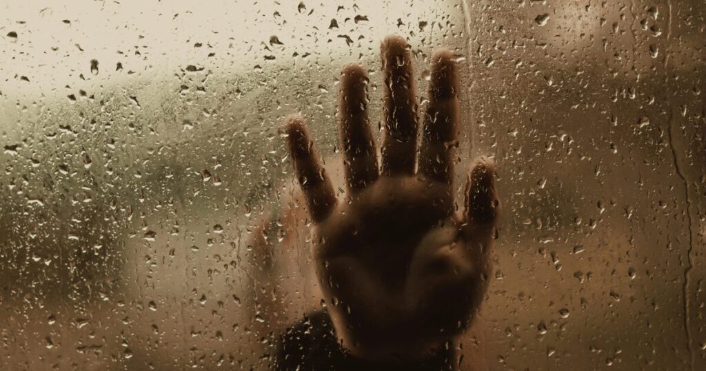 Hand pressed against a rain-splattered glass pane, water droplets blurring the view behind it.