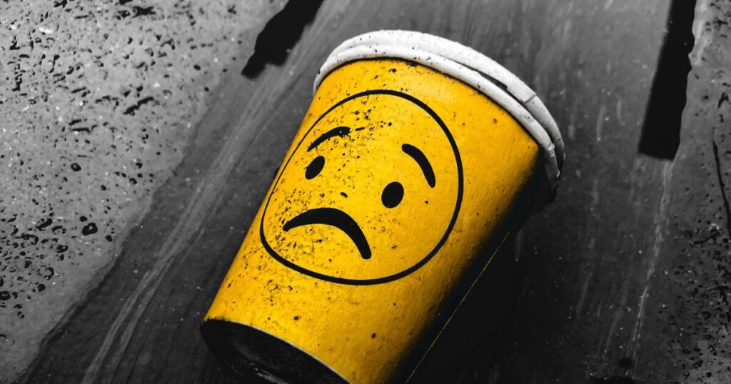 Yellow disposable coffee cup with a sad face lying on a dirty wooden surface of the floor.