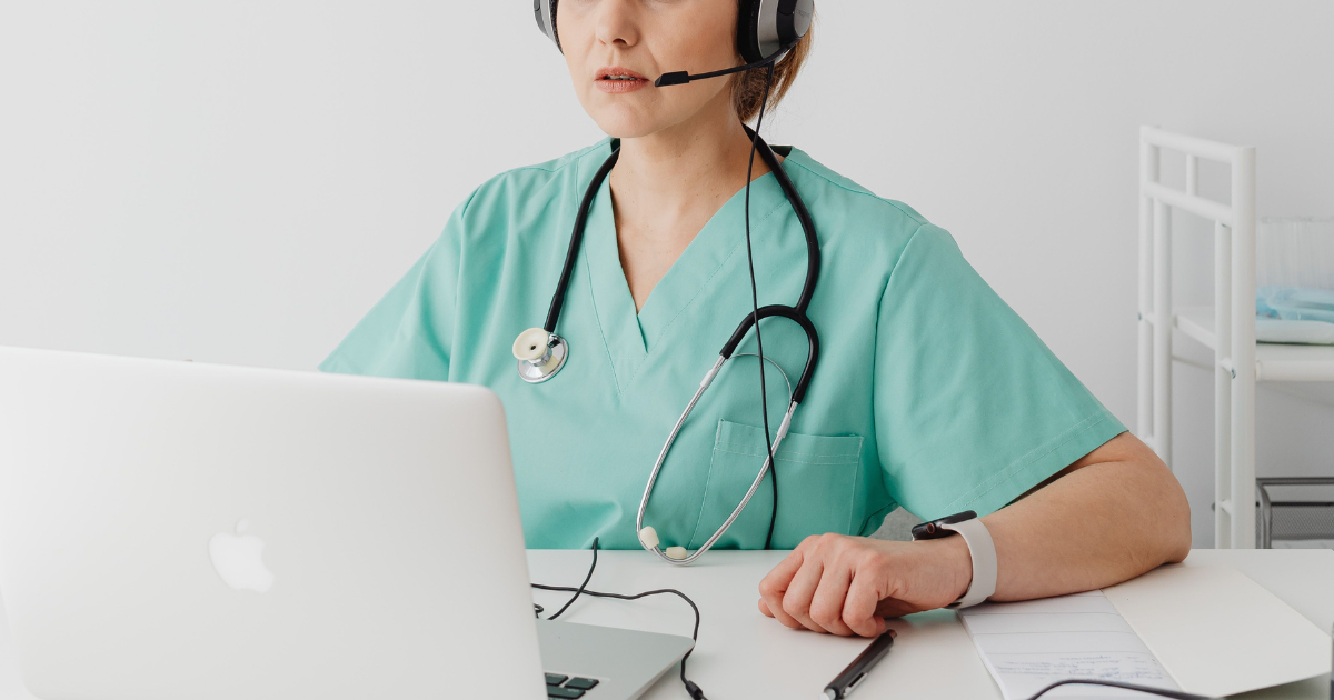 Healthcare worker wearing teal scrubs with headset and stethoscope, typing on a laptop at a desk.