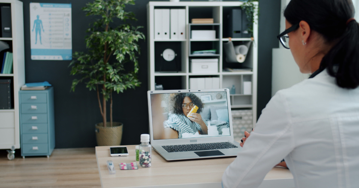 Healthcare professional at a desk during a video call; on the laptop screen a person holds a pill bottle and looks toward the camera.