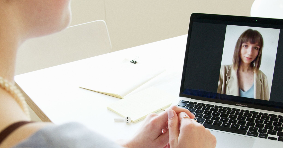 Person facing a MacBook on a white desk, video chat with a woman on screen; hands clasped, notebook and small earbuds nearby.