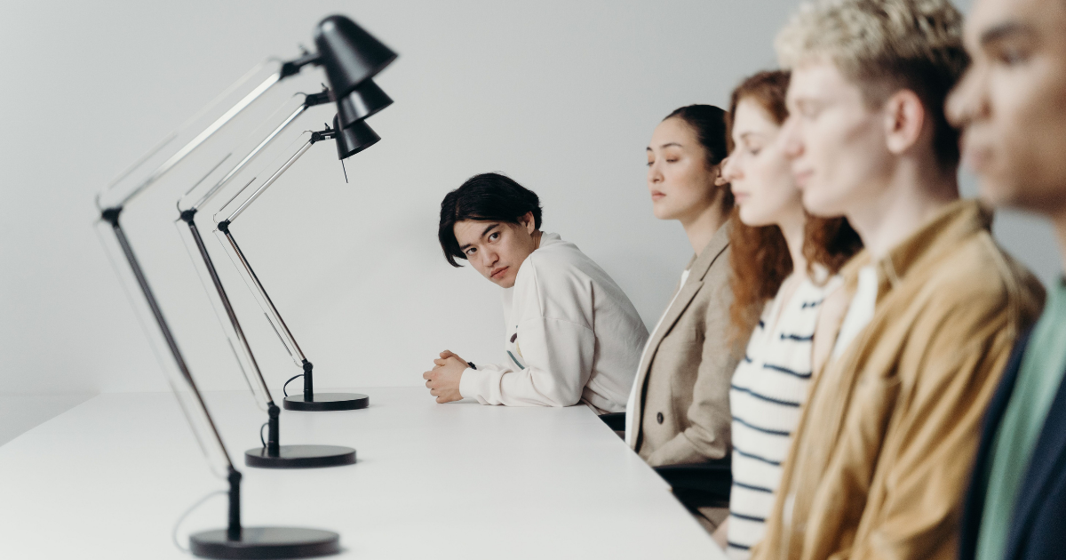 Group of five young adults seated at a white conference table with sleek desk lamps, one person glancing toward the camera.