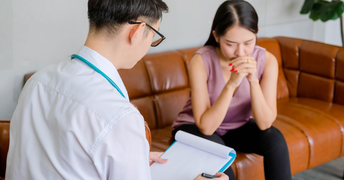 A doctor with a stethoscope reviews a clipboard as a worried patient sits on a brown leather couch, clasping her hands.