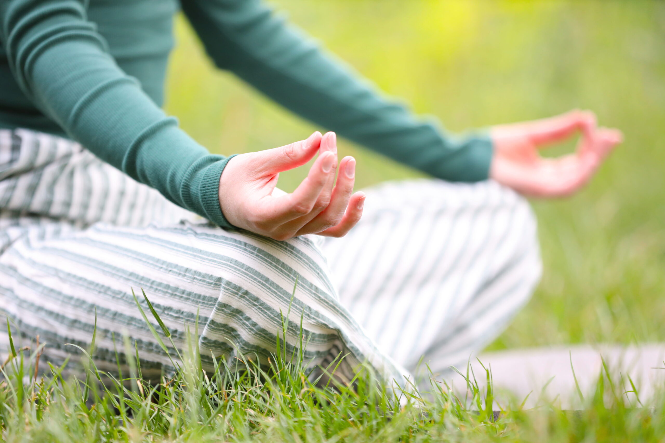 Person meditating outdoors on grass, hands in a mudra with fingers touching, wearing a green sweater and striped pants.