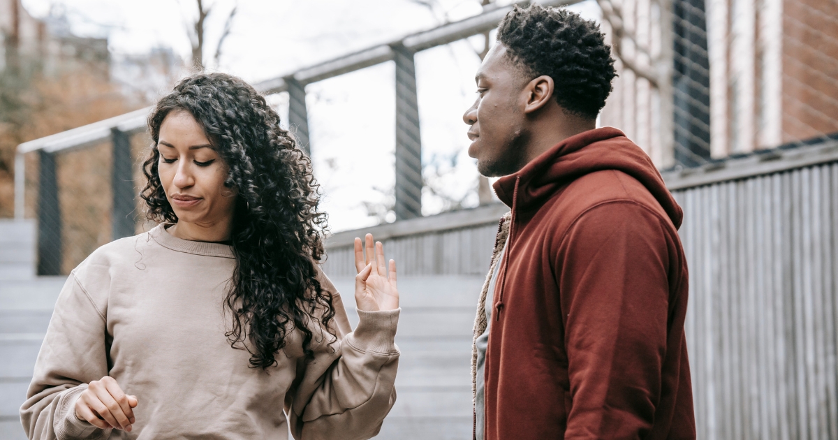 Two friends talking outdoors by a metal railing; the woman with curly hair gestures with her left hand while the man in a maroon hoodie listens nearby.