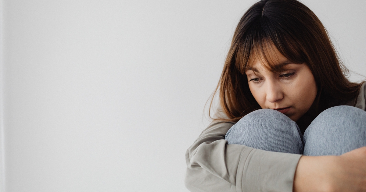 Woman sitting with knees to chest, looking down with a sad expression.