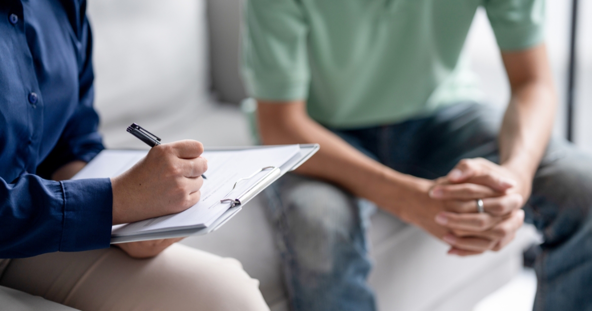 Therapist takes notes on a clipboard as a client sits nearby with hands clasped.