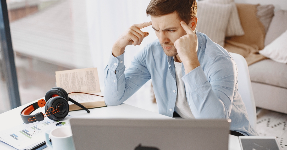 Man at a desk looking stressed, touching his temples while using a laptop and study materials at home