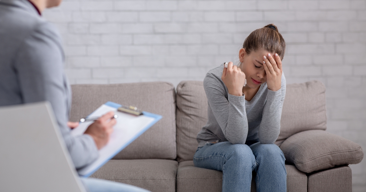 Distressed woman sits on a couch with her head in her hand during therapy, while a clinician takes notes on a clipboard beside her.