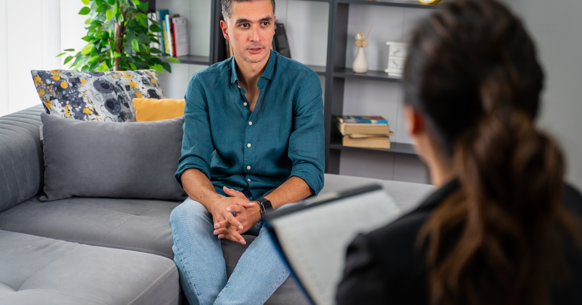 Man in a teal shirt sits on a gray sofa, hands folded, talking in a cozy living-room therapy session while a counselor notes in the foreground.