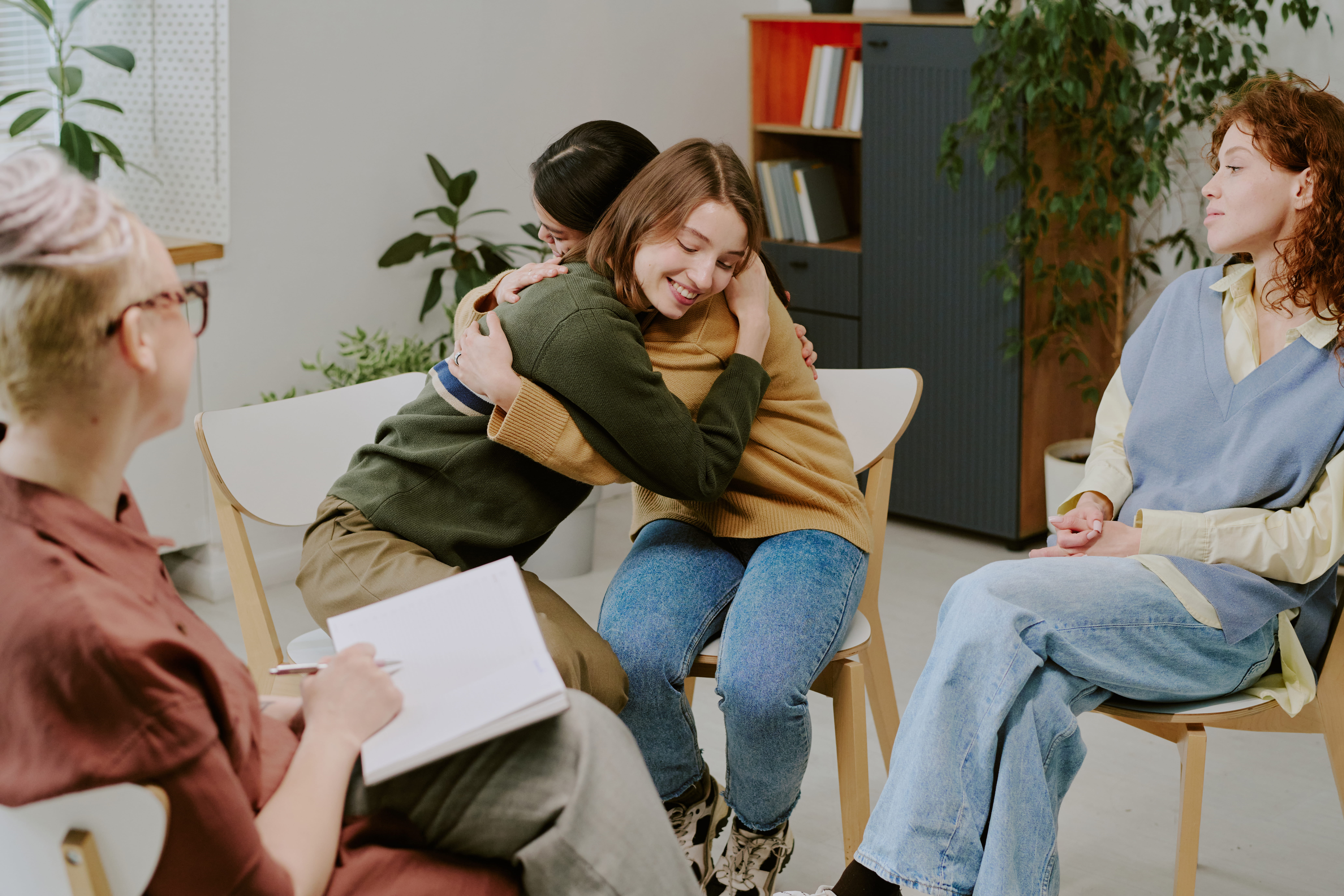 Two people share a comforting hug in a group therapy setting while others sit nearby and listen.