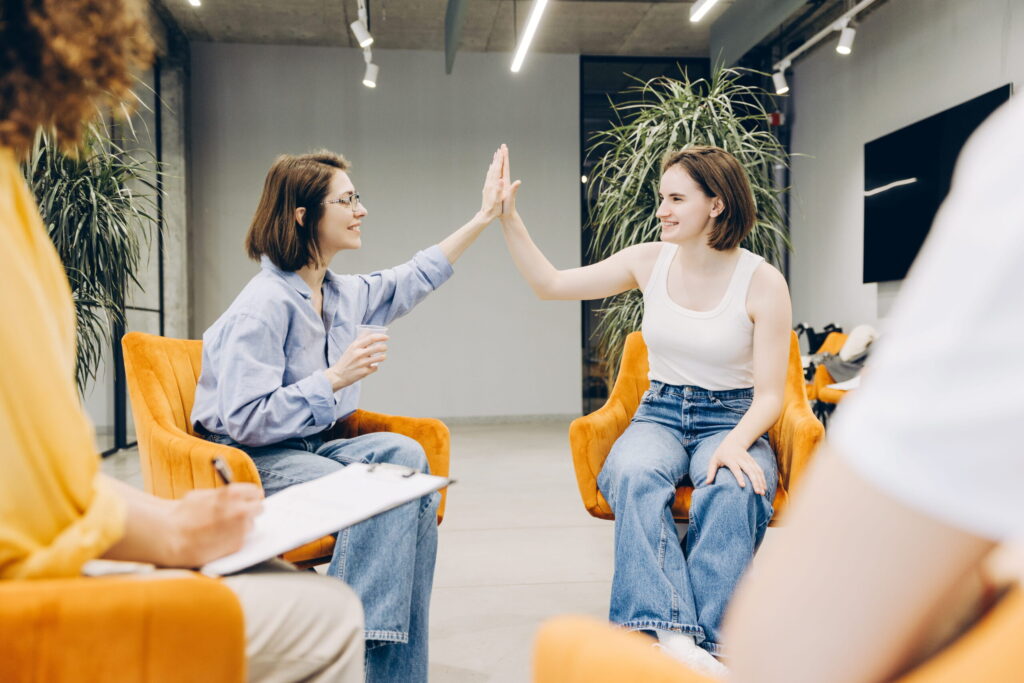 Two women sit in orange chairs and high-five in a modern office lounge.